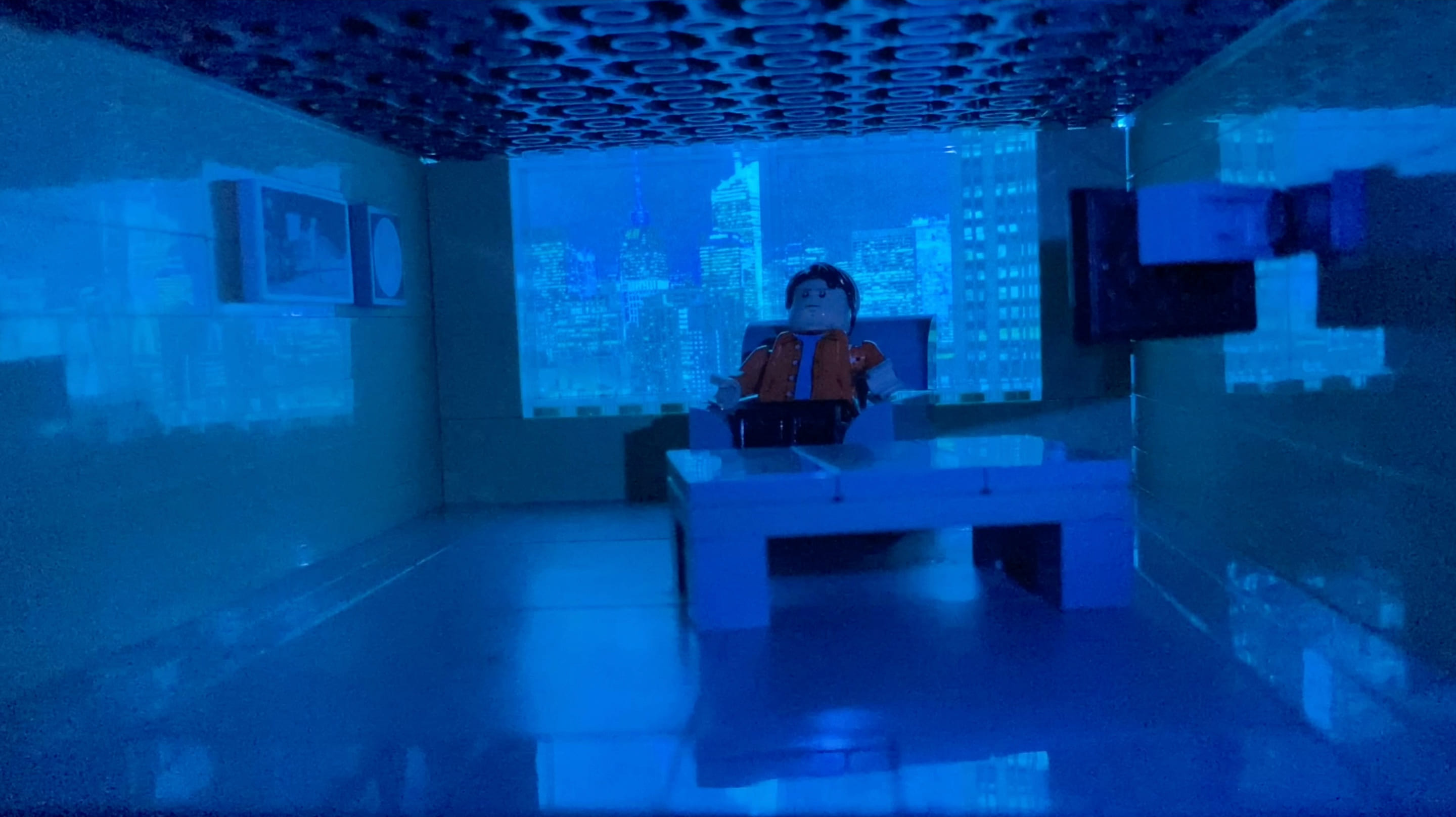 Image tirée de Back to the Moon qui est un film en Stop-Motion Lego. L’image a une teinte bleue et la figurine est assise dans un fauteuil. À gauche se trouve un tableau de la lune et de la mission Apollo 11, à droite se trouve une télévision et en face de lui se trouve une table basse grise. Derrière lui on aperçoit plusieurs gratte-ciels.